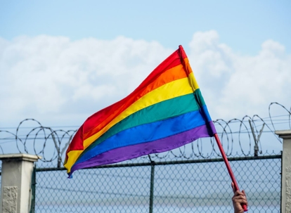 LGBTQ + flag fluttering in the breeze with a barbed wire fence in the background and a blue sky with clouds